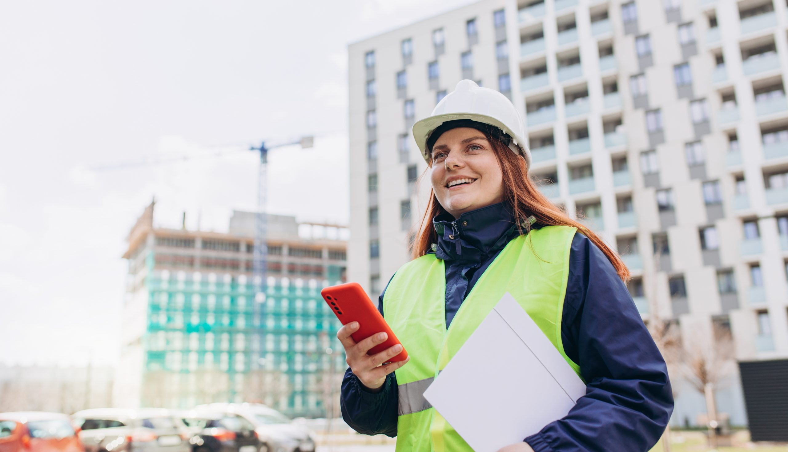 Architect with a blueprints using smartphone at a construction site. Portrait of woman constructor wearing white helmet and safety vest outdoors, technology concept